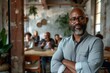 © N Joy Art  - Smiling African American man standing confidently with arms crossed in a modern office setting. Gray beard, glasses, casual yet professional clothing, colleagues in the background, radiating warmth,