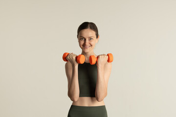 A fit woman dressed in dark activewear holds bright orange dumbbells, showcasing her enthusiasm for fitness in a simple, uncluttered environment.