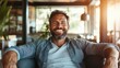 © Dulemegapixel - A cheerful bearded man sits comfortably on a couch within a sunlit room, exuding warmth and friendliness while natural light brightens the clean, modern interior.