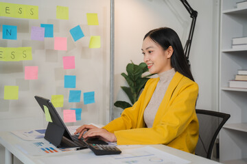 Confident Businesswoman Planning Strategy: A young, smiling Asian businesswoman works on a tablet at her desk, surrounded by colorful sticky notes and office supplies.