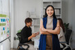 © Kritdanai - Confident Businesswoman: A professional woman stands confidently with arms crossed in a modern office setting, exuding assurance and leadership. Her colleagues work in the background.