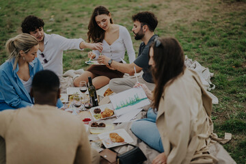 A group of friends is having a picnic while one paints in a park setting. They are surrounded by food, drinks, and enjoying a relaxed atmosphere together outdoors.