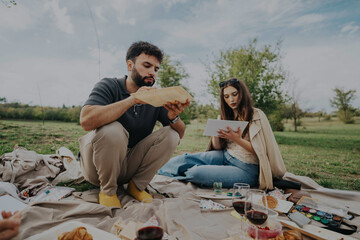 Wall Mural - A couple relaxes at a picnic with art supplies, sketching on a tablet amidst a serene park environment. Wine and snacks create a calm, creative atmosphere.