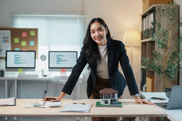 Real estate agent is leaning on her desk with a model house and paperwork, smiling confidently