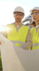 Two architects or engineers wearing safety hard hats and vests holding blueprint and discussing something on construction site at sunrise, front vertical view. Architecture engineering and teamwork