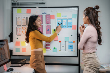 Two asian female ux ui designers are discussing over new mobile application interface design displayed on whiteboard, working together in office