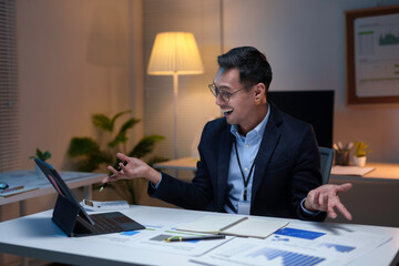 Excited businessman having a video call on a tablet and celebrating success in his office late at night