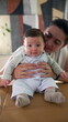 © Marco - Baby sitting on a table, supported by the mother from behind. baby looks directly at the camera with a calm expression, while the mother’s face is partially visible, creating a warm, familial moment