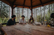 © qunica.com - A group of friends enjoying a peaceful moment in a rustic gazebo surrounded by trees. The setting features a wooden table, wine glasses, and a relaxed atmosphere, perfect for outdoor conversation.