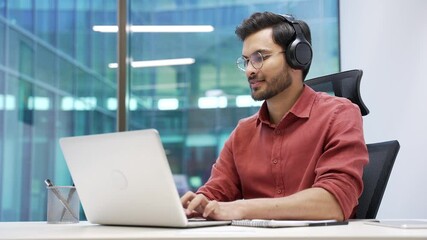 A focused IT specialist in wireless headphones works on a laptop sitting at a workplace in a business office. A handsome coder writes code. Developer is programming, busy with a project on computer
