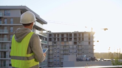 Man constructive engineer with white hard hat and safety vest is using a tablet computer while inspecting a construction site at sunset