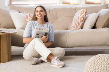 Wall Mural - Smiling young woman with magazine sitting on carpet at home