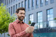 © Liubomir - Businessman smiling as he uses smartphone outside modern office building. Casual attire and positive expression suggest satisfaction, connectivity, engagement in digital communication