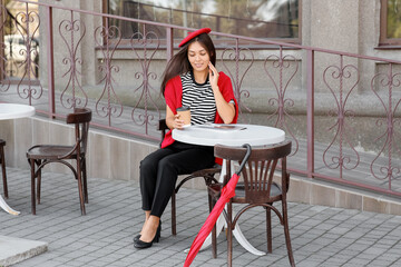 Poster - Beautiful young happy Asian woman in stylish red beret with cup of coffee sitting at city street cafe