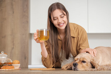 Wall Mural - Beautiful young happy woman and cute Cocker Spaniel dog with cup of green tea in kitchen at home