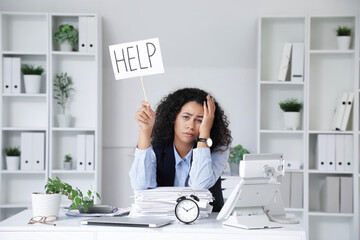 Wall Mural - Tired African-American businesswoman holding sign with word HELP at table in office. Deadline concept