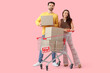 © Pixel-Shot - Happy young couple with shopping cart of cardboard boxes and FOR SALE sign on pink background