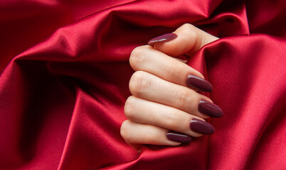 Elegant hand with burgundy nails resting on soft red satin fabric in a luxurious indoor setting