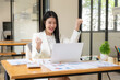 © Nittaya - Businesswoman Celebrating Success:  A young, professional woman  in a modern office setting,  exudes joy and excitement as she celebrates a successful achievement on her laptop.