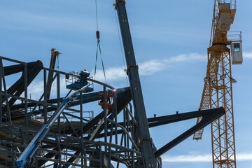 Construction workers on a crane assembling a steel structure under a clear blue sky