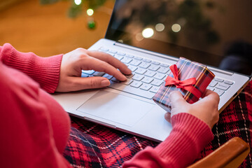 Christmas. Woman using laptop for searching gift ideas sitting at table with ready gift boxes and cup of cocoa and marshmallows near fireplace and christmas tree. Concept