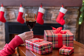 Happy woman in red christmas hat covered plaid with glass of wine sitting and relaxation on armchair near fareplace and fir-tree after finishing pakking gift boxes for family.