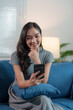 © crizzystudio - A happy attractive Asian woman sits on a sofa in the living room using a mobile phone connected to the internet to play games. online shopping  View social media, online chat, technology concept