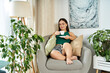 © LIGHTFIELD STUDIOS - A young brunette woman with short stature sits comfortably in her cozy living space, savoring a warm drink while surrounded by greenery.
