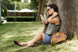 © LIGHTFIELD STUDIOS - A carefree brunette woman enjoys her time outdoors, leaning back against a tree, absorbed in her smartphone.