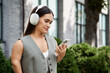 © LIGHTFIELD STUDIOS - A young brunette woman with headphones smiles while looking at her smartphone in a vibrant outdoor setting.