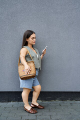 A young woman with short stature stands outside, absorbed in her phone, dressed in casual summer attire with a woven bag.