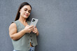 © LIGHTFIELD STUDIOS - A cheerful brunette woman stands against a wall, engaging with her smartphone on a bright, beautiful day outside.