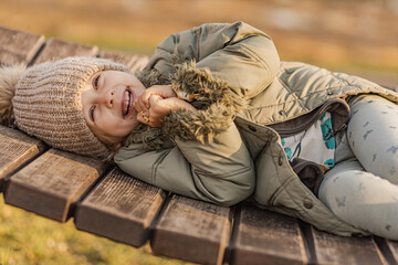 A young girl wearing beige knit cap, is lying on a wooden bench and laughing, her face partly covered by her hand. Laughing child in autumn park. Outdoor activities