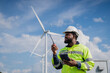 © VStudio - A man in a yellow safety vest is holding a walkie talkie and looking up at a wind turbine. electrical, engineer, environment, energy, clean, sustainable, wind turbine, nature