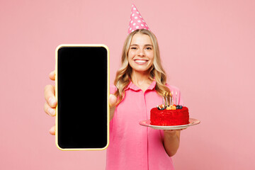 Young smiling happy woman wears shirt cone hat celebrating birthday holiday hold in hand cake use show blank screen area mobile cell phone isolated on plain pastel pink background. Lifestyle concept.