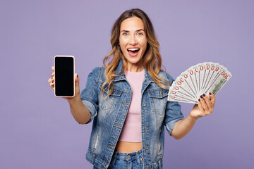 Young woman wears denim jacket violet top casual clothes hold use blank screen mobile cell phone, fan of cash money in dollar banknotes isolated on plain pastel purple background. Lifestyle concept.