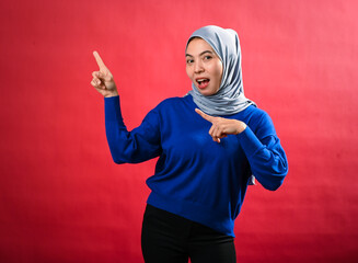 An Asian woman wearing a grey hijab and blue sweater points enthusiastically in two directions with both hands. She smiles playfully while standing against a vibrant red background