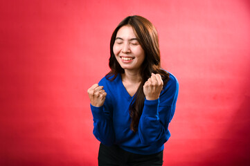 An Asian woman in a blue sweater and black pants stands against a red background with her eyes closed, fists clenched, and an excited expression, as if celebrating a victory.