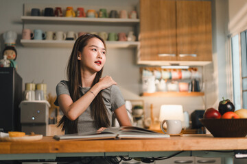 young woman sits at wooden table in cozy kitchen, looking thoughtfully at book. warm lighting and colorful mugs in background create welcoming atmosphere