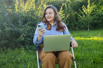 Freelance concept. Positive young woman in stylish outfit using notebook and messaging on smartphone while working on project on laptop in nature outdoors. People urban lifestyle concept. High quality