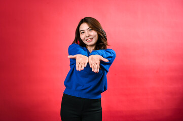 An Asian woman, wavy brown hair smiles warmly as she extends both hands forward, palms up, in a welcoming gesture. wearing a blue sweater, standing against a red background