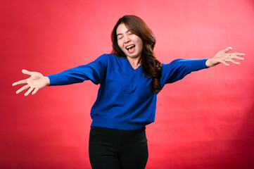 An Asian woman with long, wavy brown hair joyfully poses with open arms in a vibrant studio shot. She is wearing a blue sweater and black pants, smiling widely against a bright red background