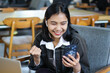 © M Alfan Setyawan - excited asian woman holding mobile phone and sitting in cafe with laptop celebrate victory by raising fist pump show her succeed