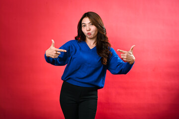 Asian woman standing against a red background, wearing a blue sweater and black pants. She is smiling and pointing at herself with both hands, showcasing a cheerful and confident expression