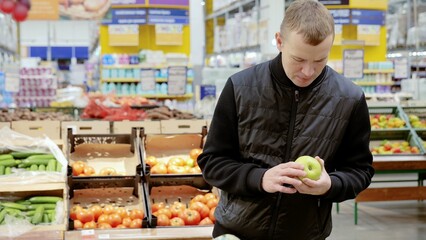 Customer carefully choosing a green apple in a grocery store produce section with vibrant fruits and vegetables in the background. Blurred shelves add freshness and variety