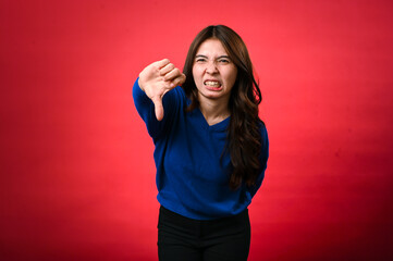 An Asian woman in a blue sweater showing a displeased facial expression, giving a thumbs-down gesture with her right hand. The background is red