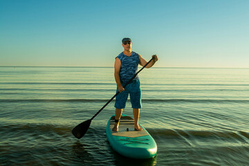 A man in sports shorts and a T-shirt on a SUP board with a paddle in a calm sea in the morning