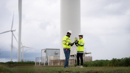 two workers in yellow safety vests are standing next to a large wind turbine. they are looking at a 