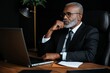 © Anastasiya - A thoughtful senior businessman in a black suit, sitting at his desk with a laptop, deep in thought while working in a modern office setting, conveying professionalism and focus