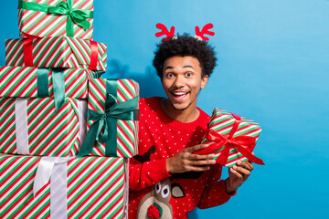 Photo portrait of attractive young man hide pile gifts hold present dressed red christmas sweater theme party on blue color background